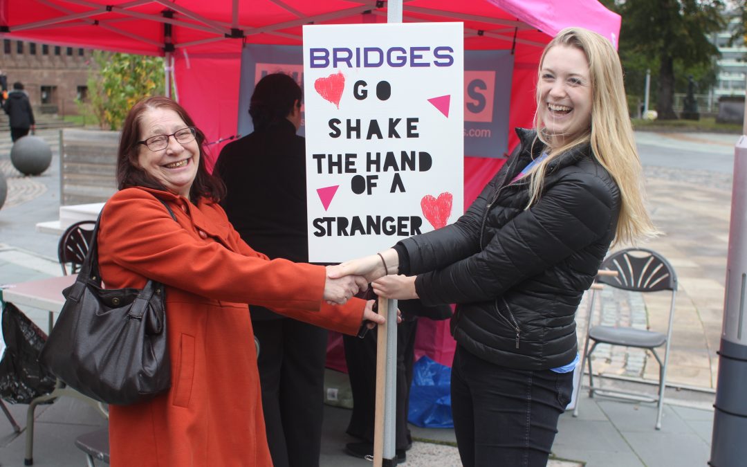 A stall set up in public for the positive placards project, with two women shaking hands alongside a picket board stating "Bridges, go shake the hand of a stranger"