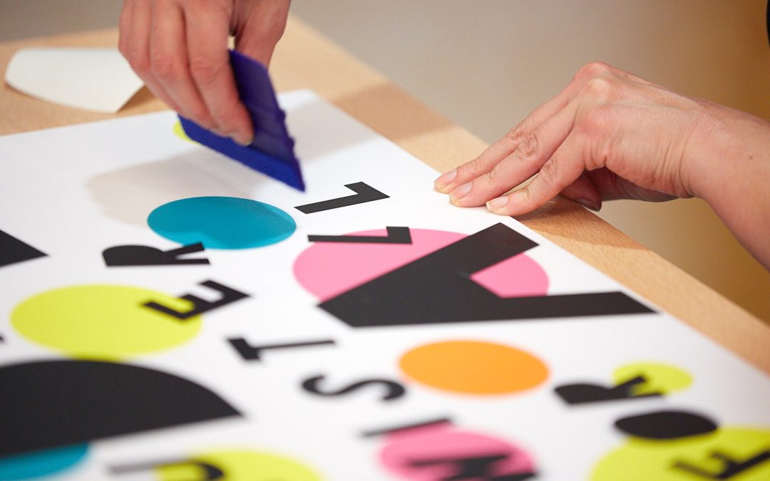 A person gluing cut out black letters and blue, orange and yellow circles to create a poster.
