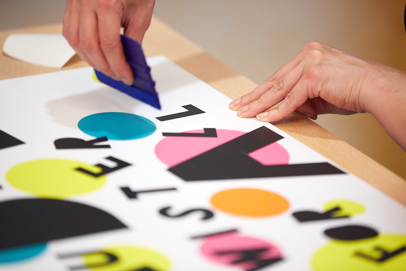 A person gluing cut out black letters and blue, orange and yellow circles to create a poster.