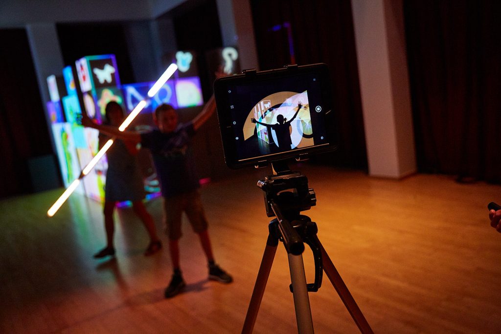 Illuminated cubes in background with silhouettes of two people, one of them is holding a long LED tube light. You can see a reflection of all of this on the camera screen in the foreground, as well as the camera set up.
