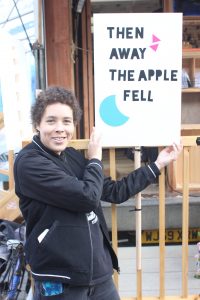 A lady standing beside a sign reading "Then away the apple fell"