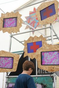 A boy is using an iPad to control geometric visuals on 5 wooden framed monitors mounted to a scaffolding tower.